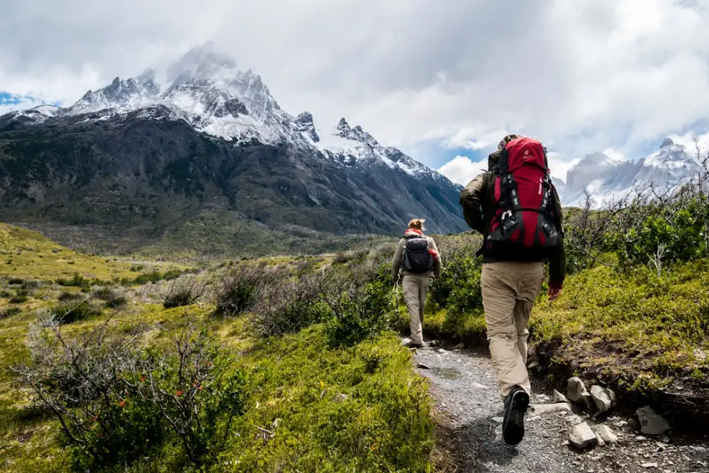 Two hikers with backpacks walking along a trail toward snow-capped mountains