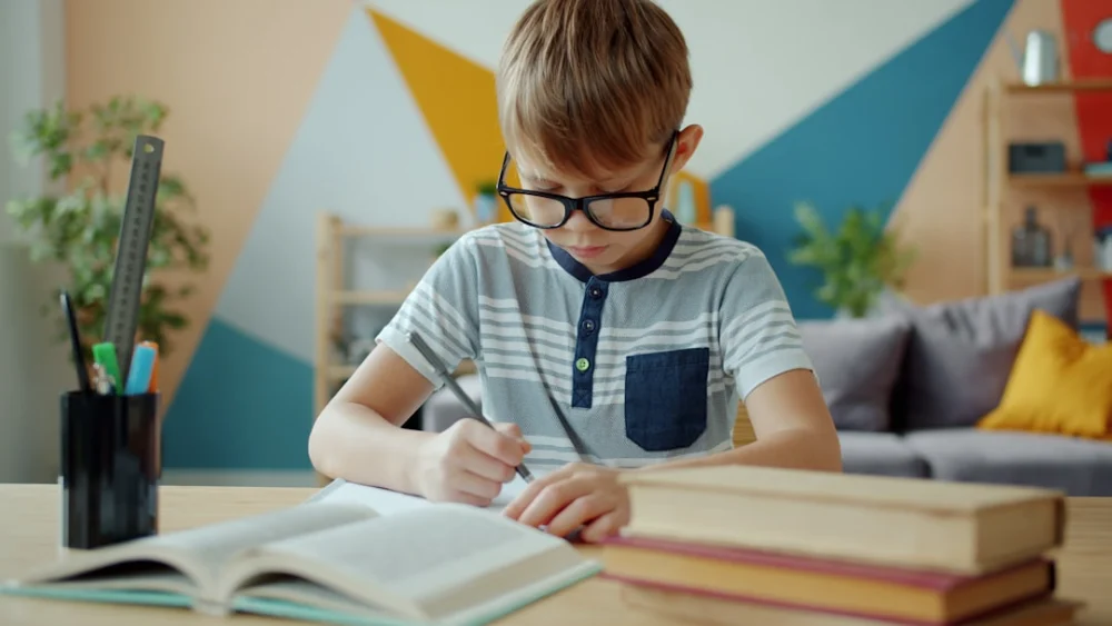 Young boy with glasses writing at desk with books