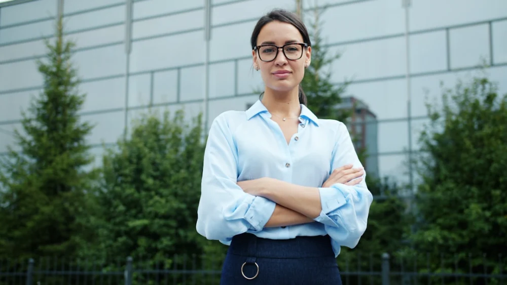 A woman in glasses stands with arms crossed outdoors