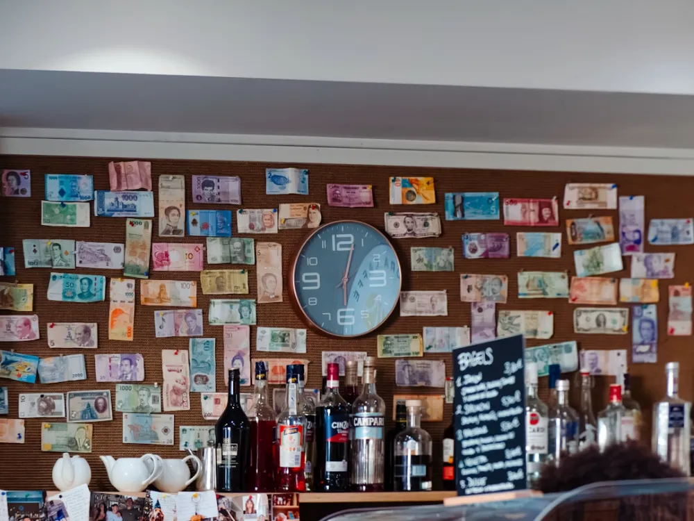 A wall covered in international banknotes with a round clock and liquor bottles