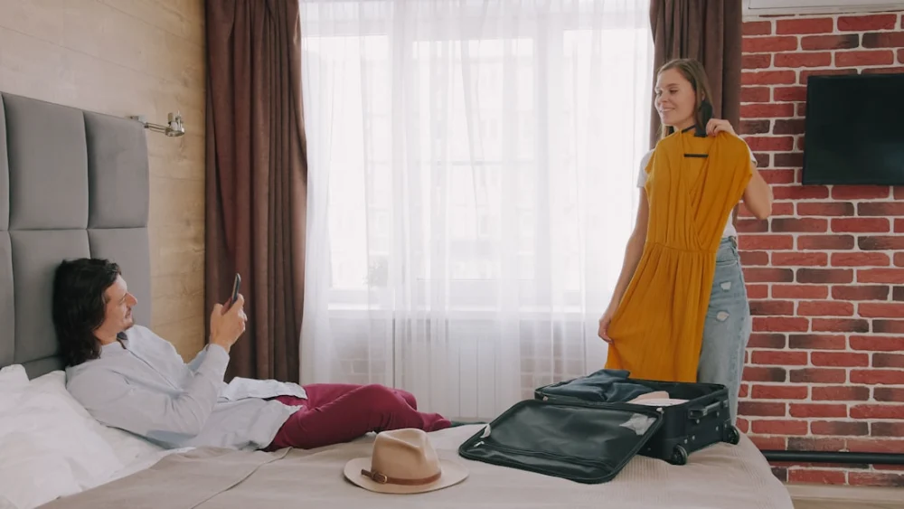 A woman holding a yellow dress while a man uses a phone on a bed
